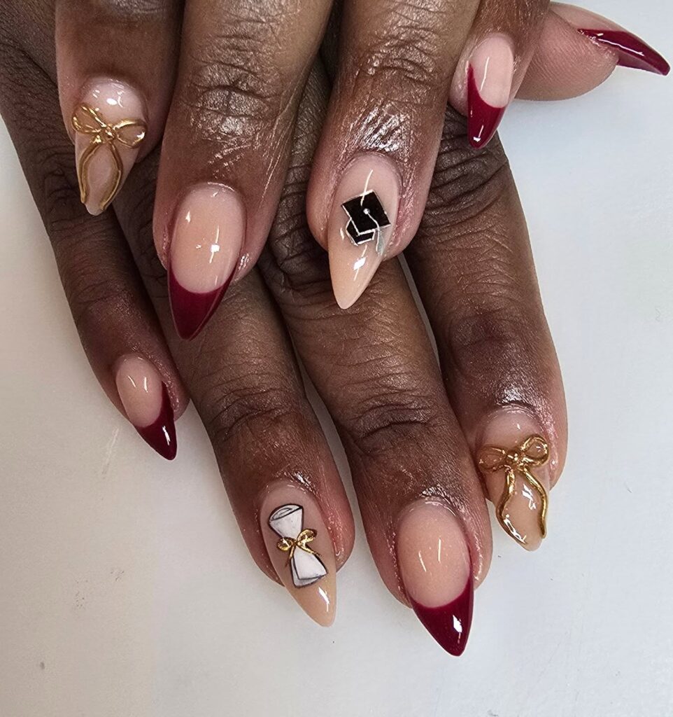 Stiletto Nails Featuring Warm Nude and Deep Burgundy French Tips with 3D Gold Bows and Graduation Cap Accents